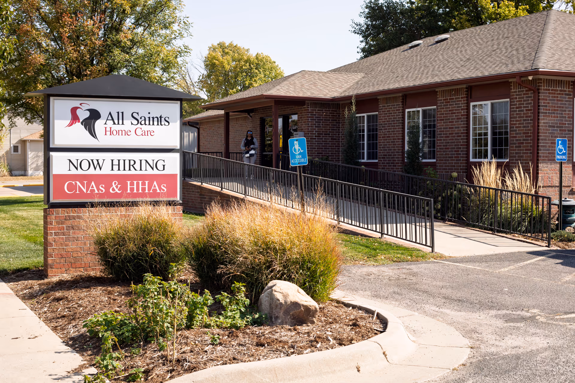 Exterior view of All Saints Home Care building with a Now Hiring sign and wheelchair ramp.