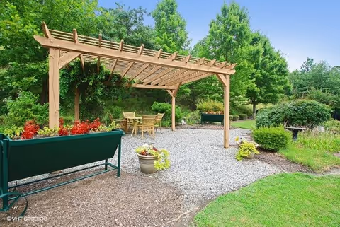 Outdoor garden area with a wooden pergola over a gravel patio. Under the pergola, there is a table with four chairs. The surrounding area has green grass, bushes, trees, and raised garden beds with flowers and plants.