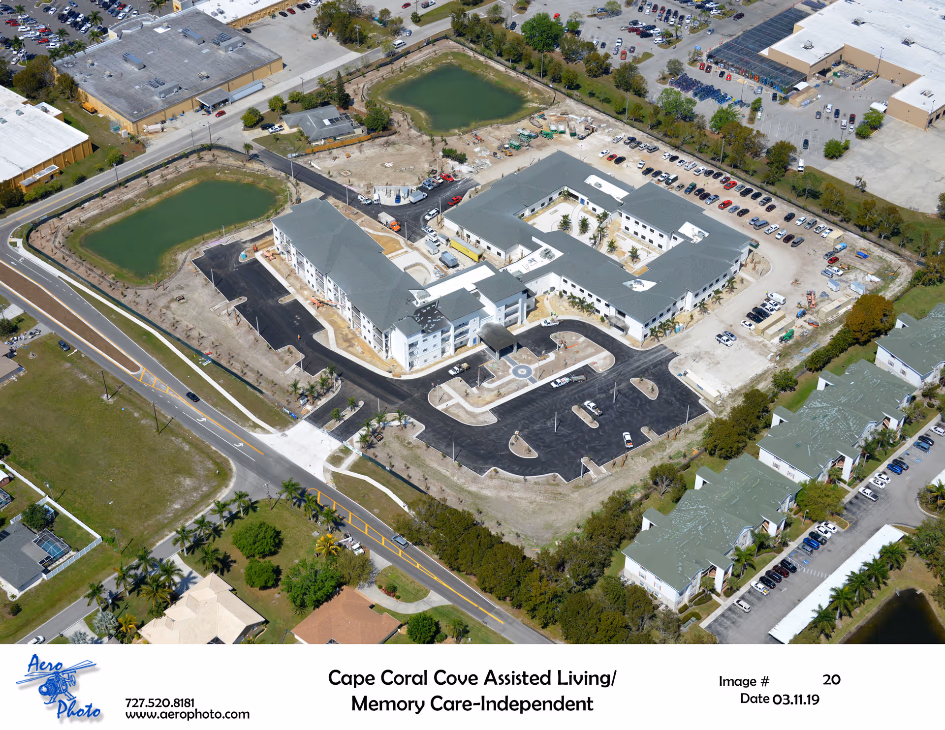 Aerial view of Cape Coral Cove Assisted Living and Memory Care-Independent facility showing multiple buildings with gray roofs, parking lots, surrounding roads, and two small ponds nearby.