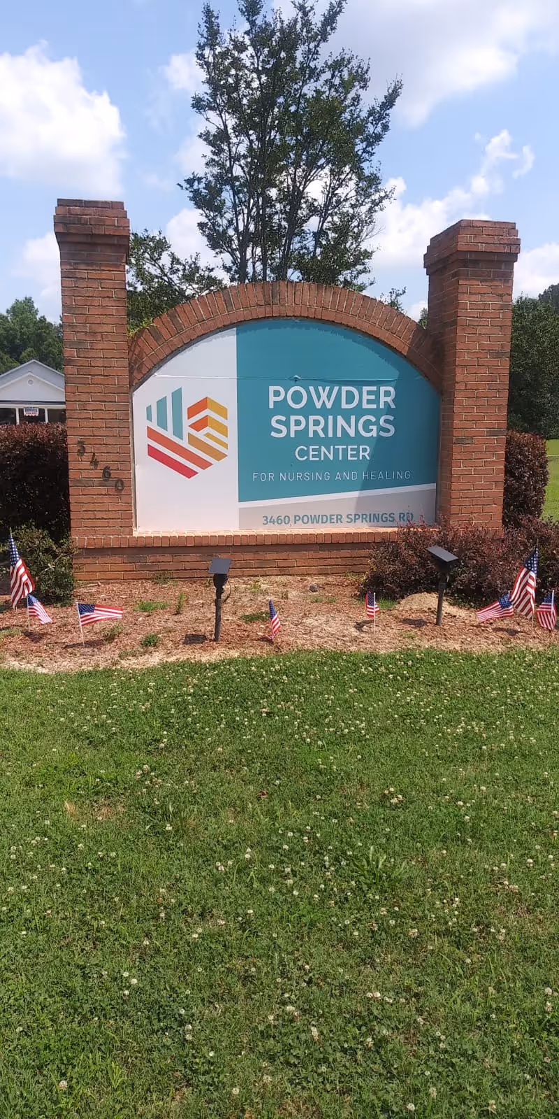 Brick entrance sign that reads 'Powder Springs Center for Nursing and Healing' with small American flags in the landscaped foreground and grass in front.