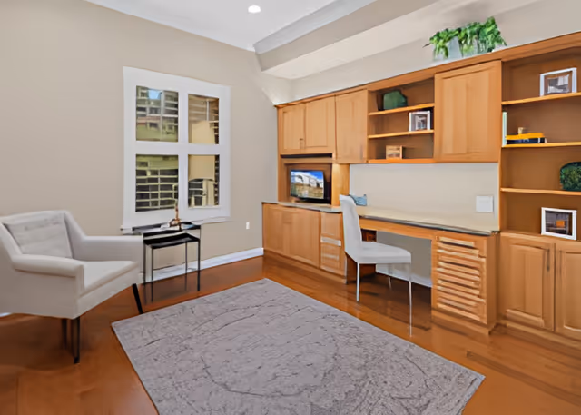 Bright furnished room with built-in wooden desk and shelving, a chair and armchair, window and area rug.