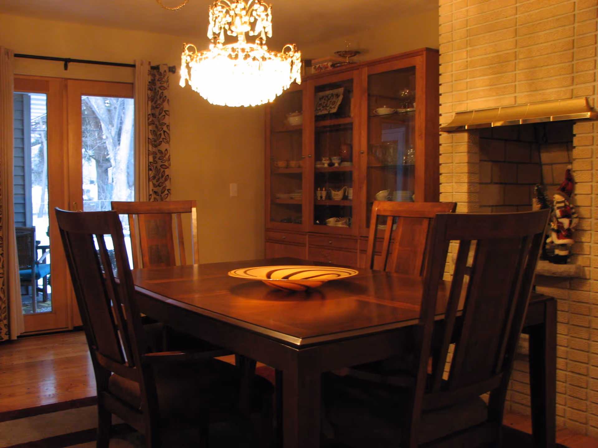 Wooden dining table with chairs and a decorative bowl under a chandelier in a warmly lit dining room with a china cabinet and glass doors.