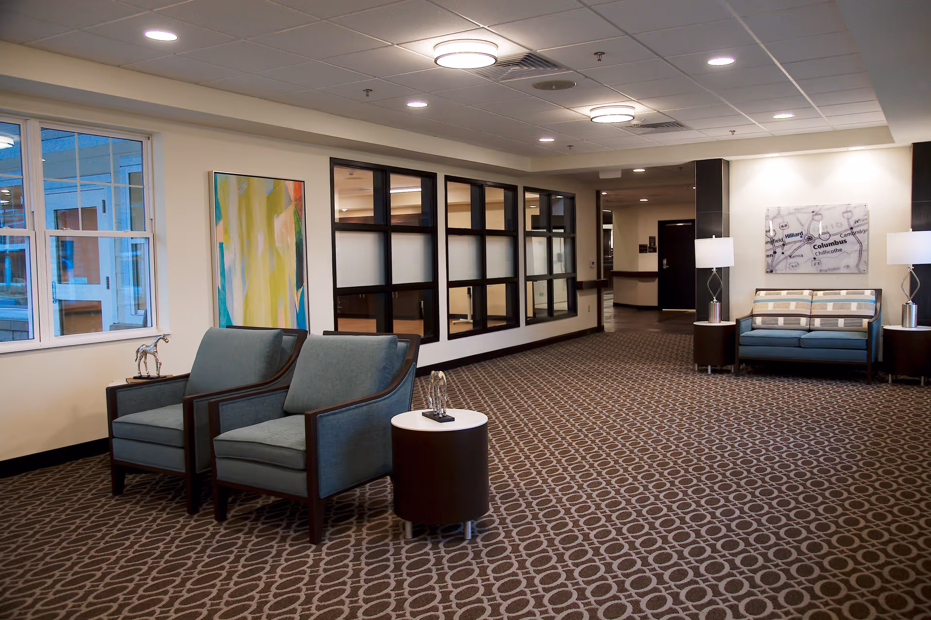 A spacious and well-lit common area with patterned carpet flooring, two blue upholstered armchairs with wooden frames, a small round table between them, and a blue upholstered loveseat against the far wall. The walls are decorated with abstract artwork and a map. There are two table lamps on either side of the loveseat, and large windows on the left side letting in natural light.