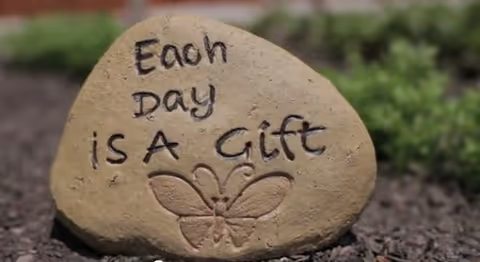 A decorative garden stone engraved with 'Each Day is A Gift' and a butterfly, resting on mulch with greenery in the background.