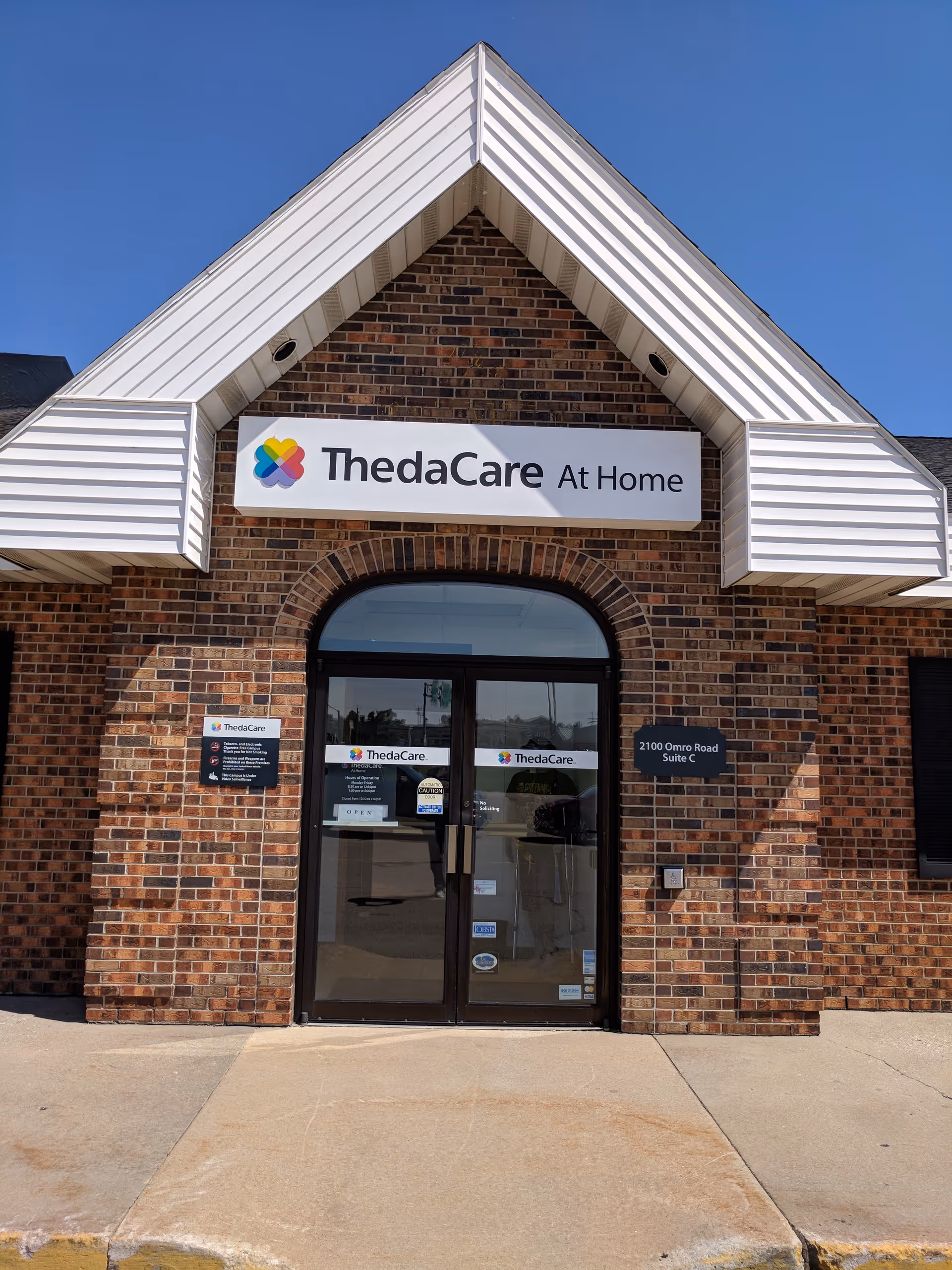 Brick building entrance with ThedaCare At Home sign and glass doors.