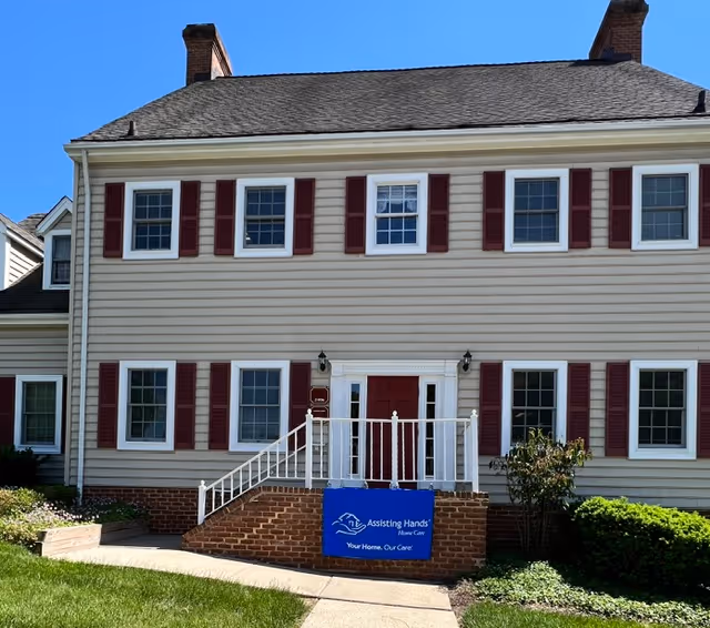 Two-story beige building with red shutters, white railings, stairs, and a blue Assisting Hands banner.