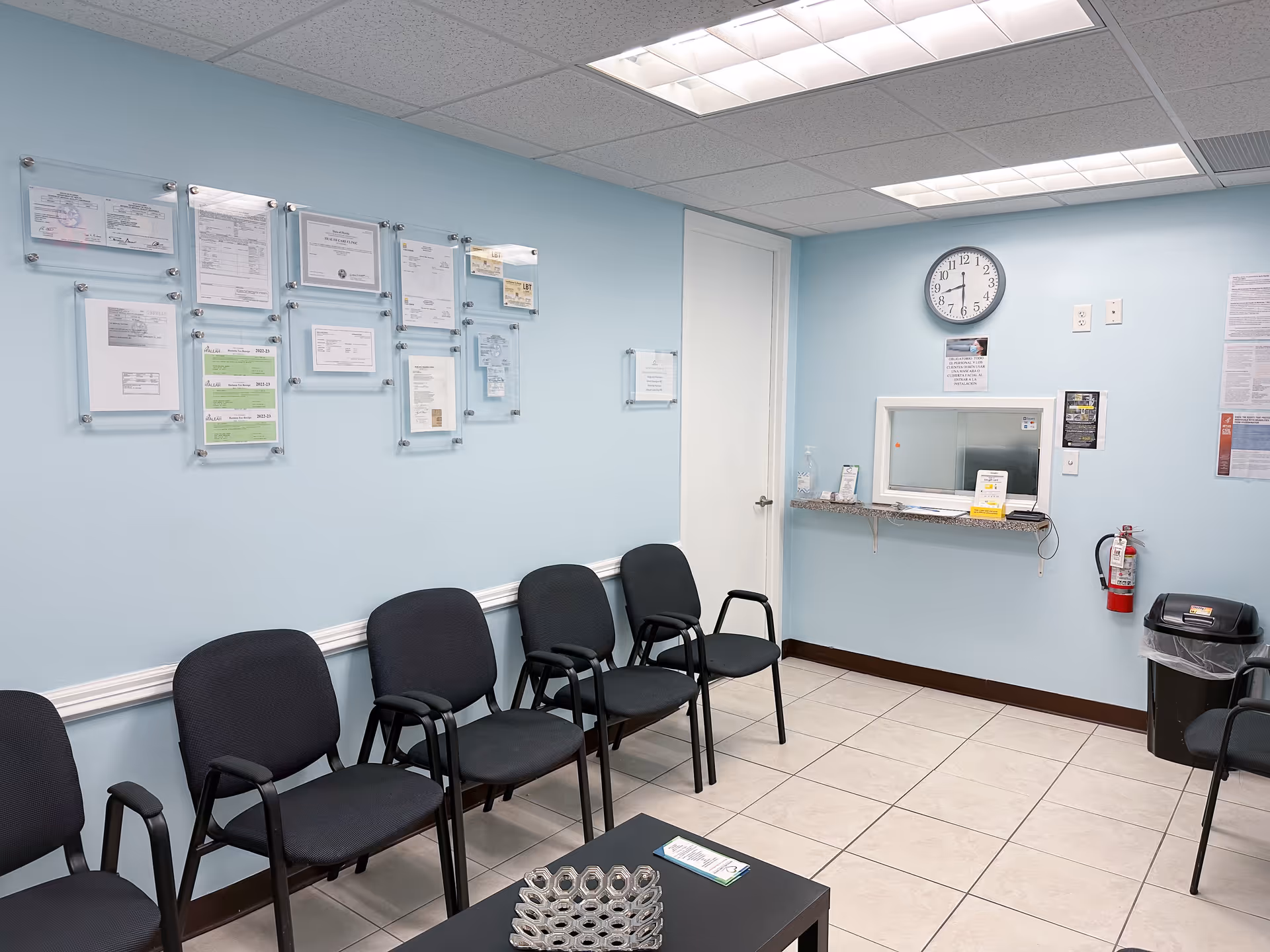Row of black chairs along a pale blue wall with a reception window and clock.