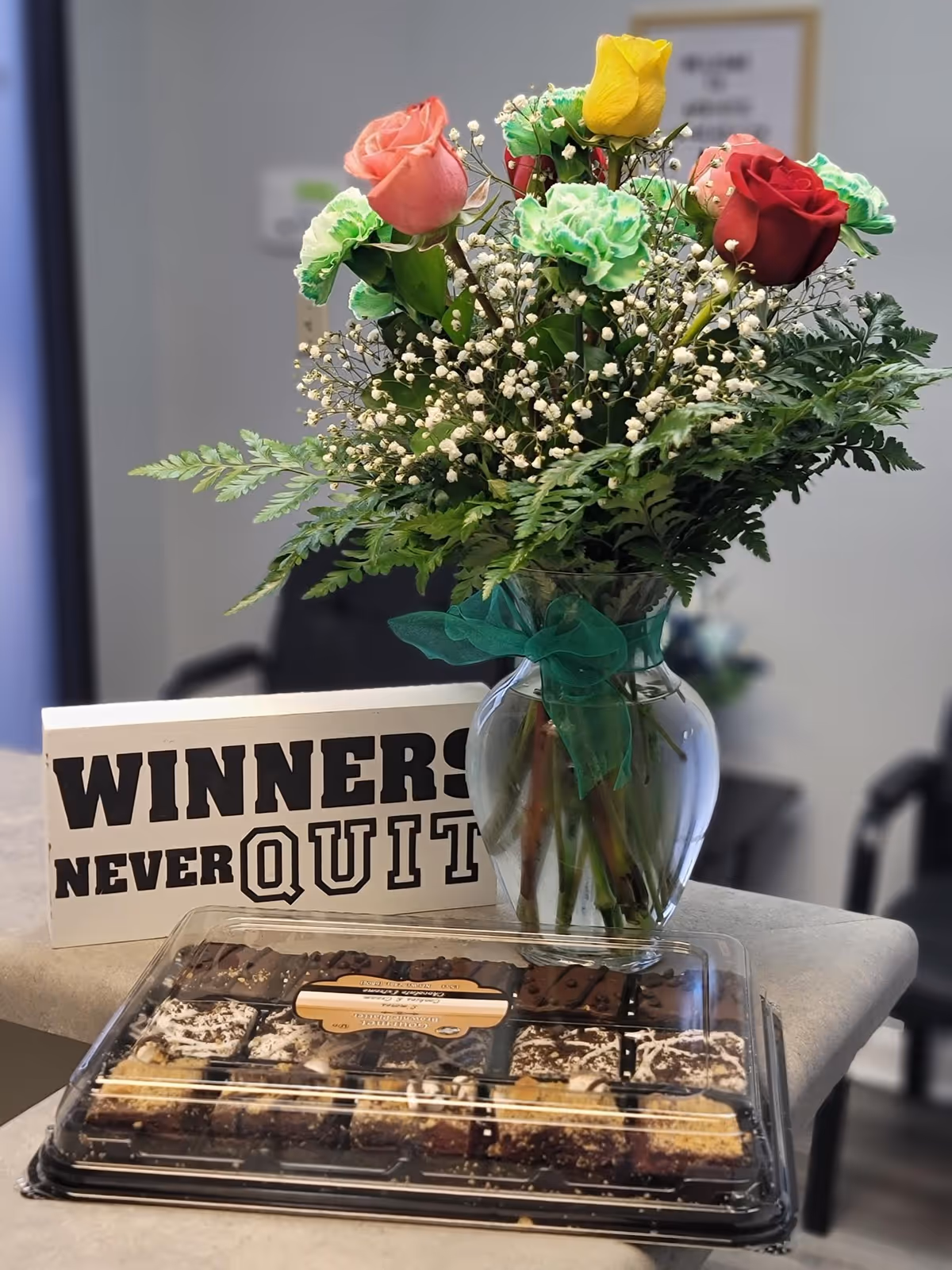 Bouquet in a glass vase with a 'Winners Never Quit' sign and a chocolate tray.