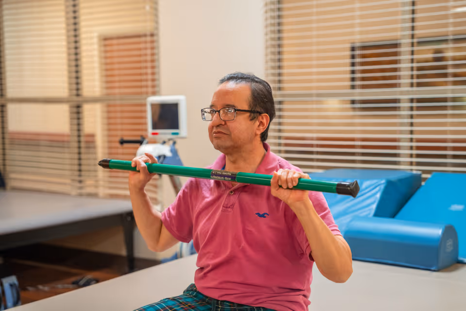 A man wearing glasses and a pink polo shirt is sitting indoors holding a green exercise bar across his shoulders. Behind him are blue padded exercise mats and a window with horizontal blinds.