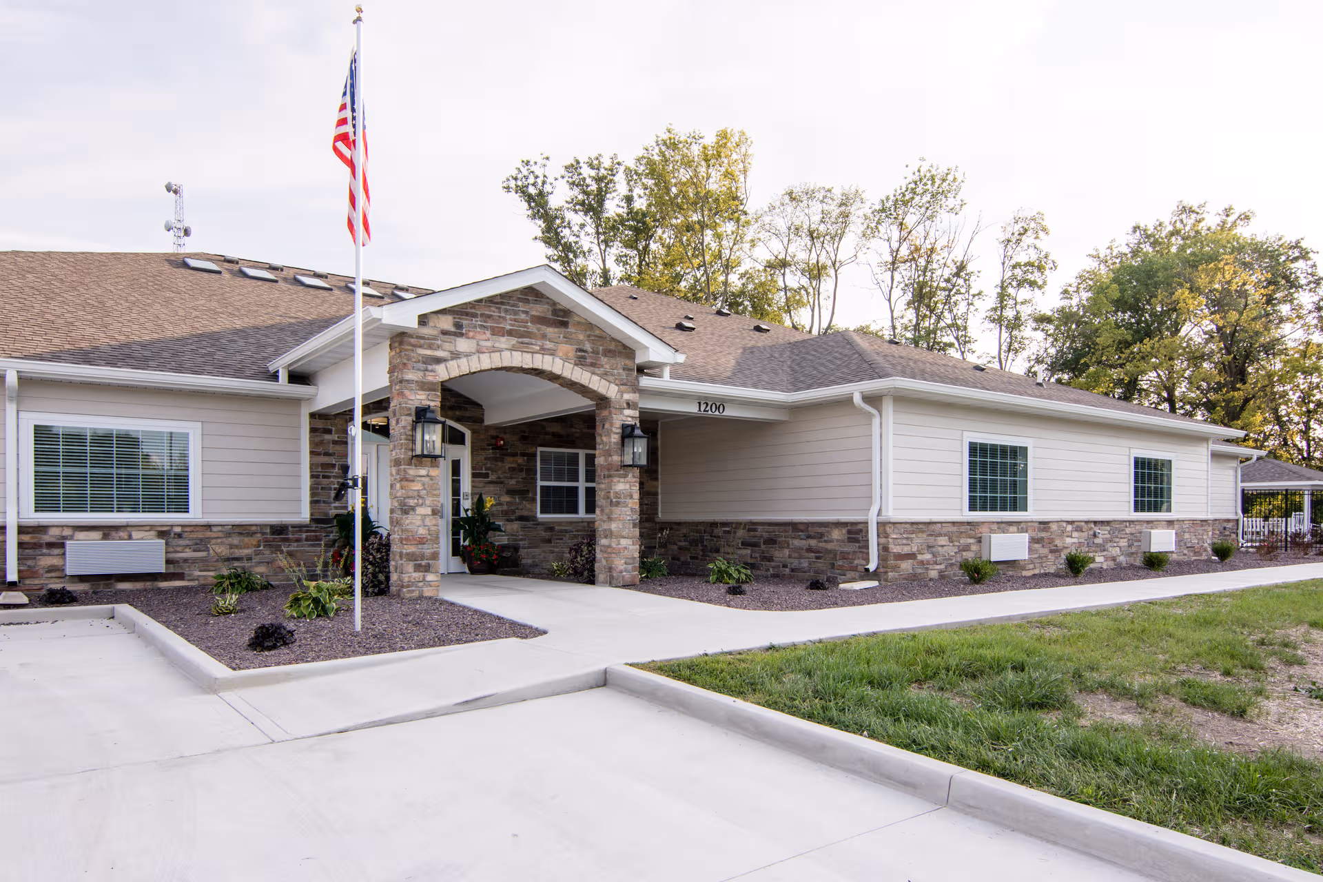 Single-story memory care facility exterior with stone facade, arched entrance, flagpole, and landscaping.