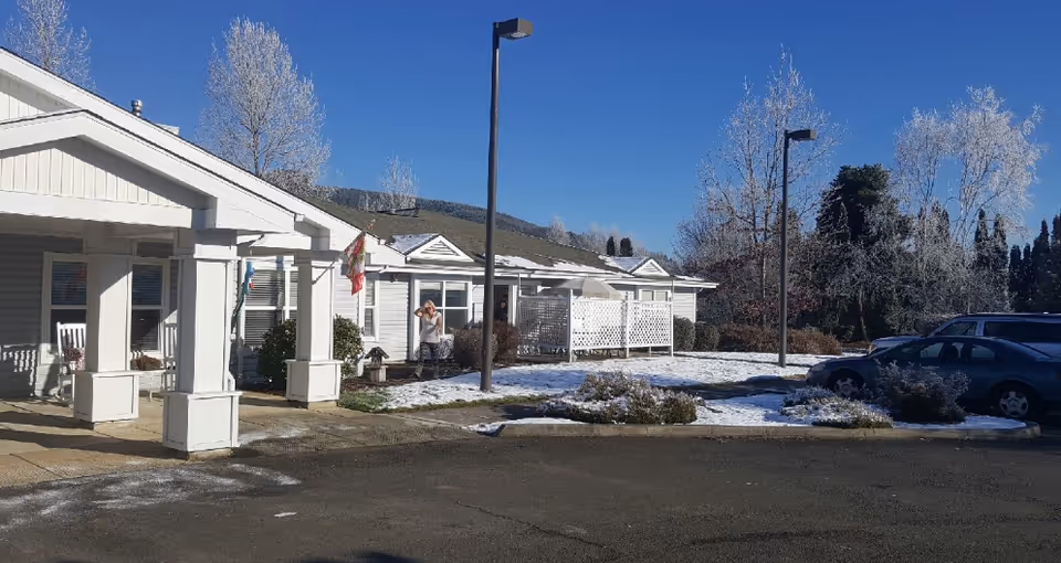 White single-story Applegate Place exterior with a covered entrance, flags, cars, and light snow.