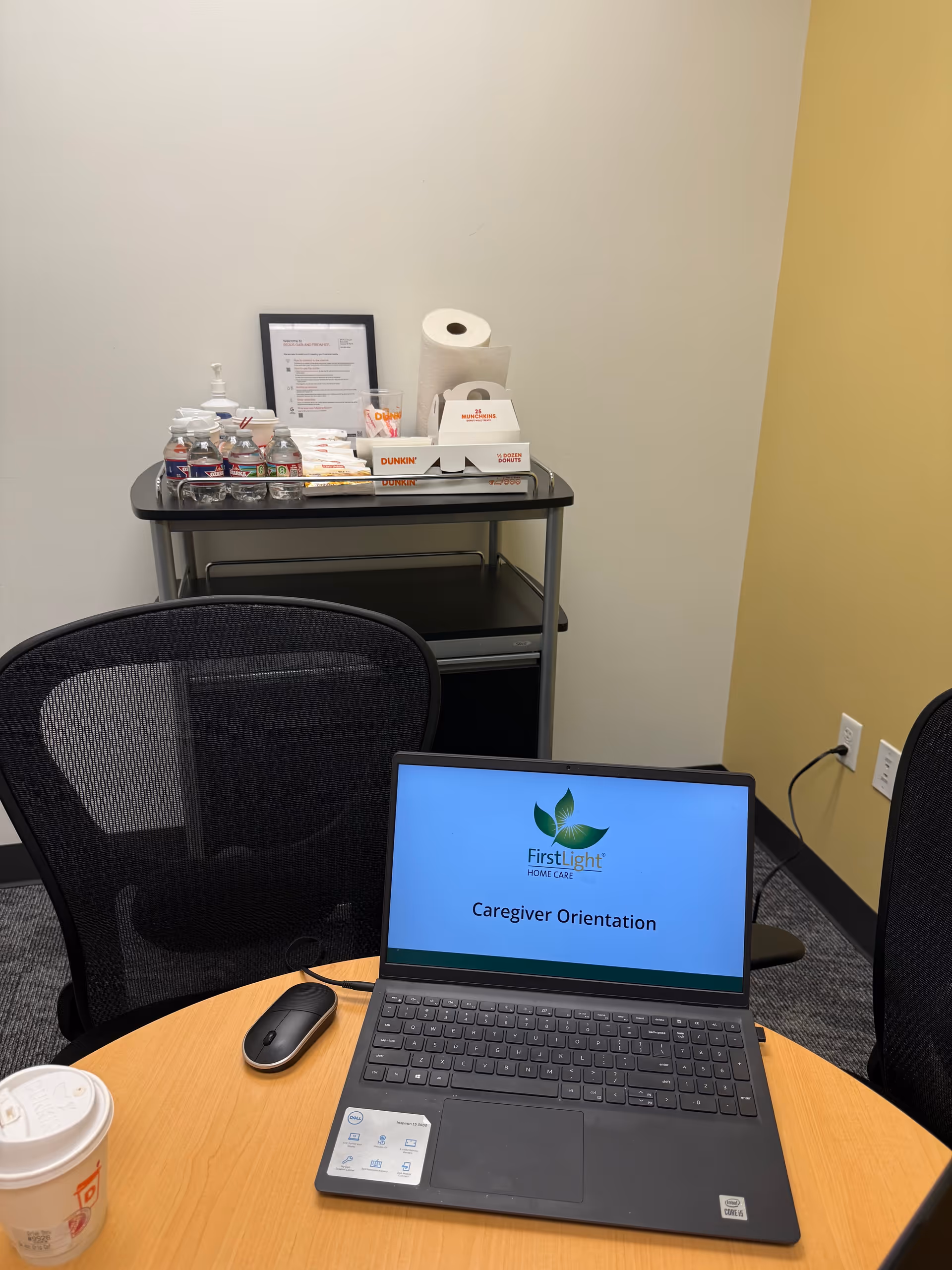 Laptop on round table displays Caregiver Orientation; supplies cart and coffee cup nearby.