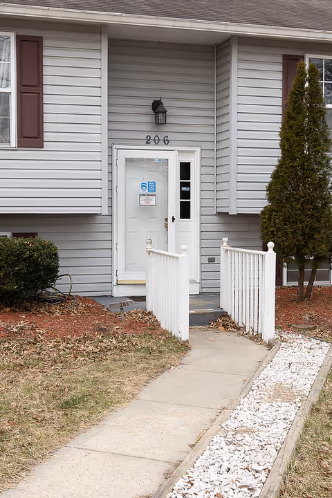 Entrance to a residential building with gray siding and maroon shutters. The door is white with the number 206 above it. A concrete walkway leads to the door, flanked by white railings and a small landscaped area with mulch, bushes, and a tree.