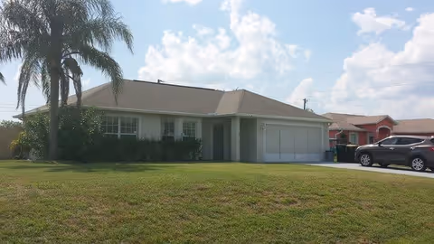 Single-story house with a front lawn, palm trees, and a driveway with a parked car under a partly cloudy sky.