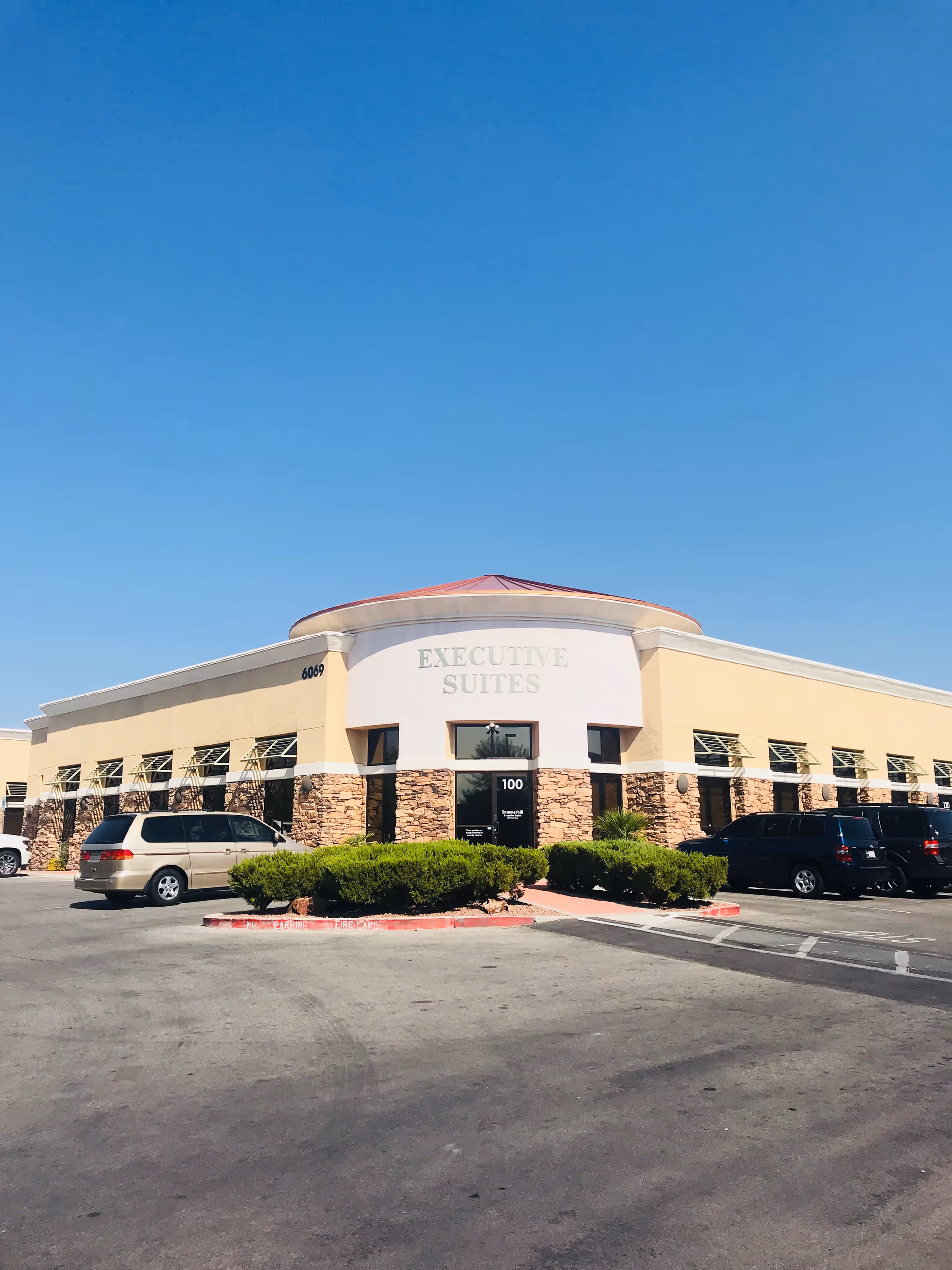 Sunny exterior of a beige building with stone accents, Executive Suites sign, and parked cars.