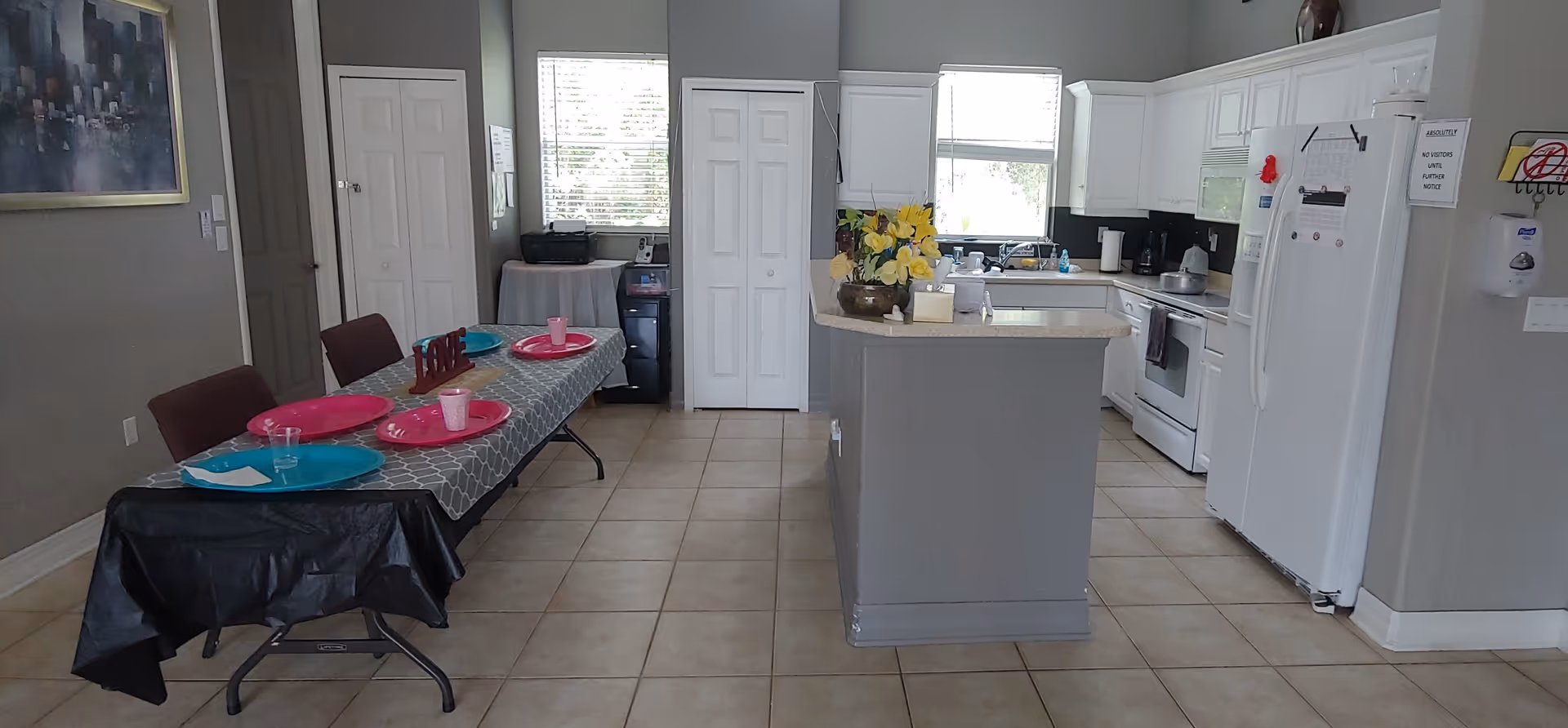 A bright kitchen and dining area with tiled floors. The kitchen features white cabinets, a white refrigerator, stove, and microwave. A kitchen island with a flower arrangement is in the center. To the left, a dining table is set with colorful plates and cups, and chairs are arranged around it. Two windows provide natural light, and there is a painting on the left wall.