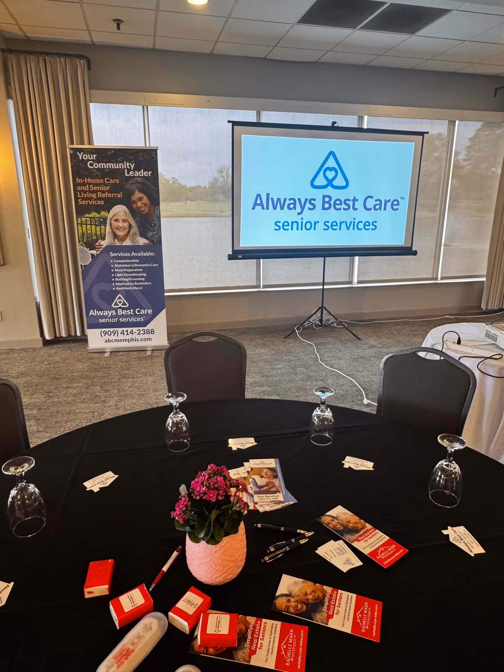 Round table with brochures, banners, and a projector screen displaying Always Best Care logo.