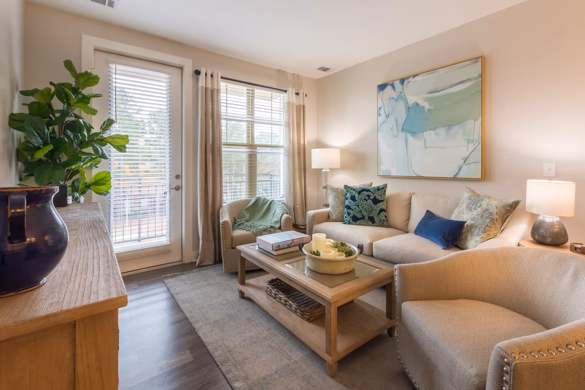 A cozy living room with a beige sofa adorned with decorative pillows, two beige armchairs, a wooden coffee table with candles and books, a large abstract painting on the wall, a wooden sideboard with a large blue vase and a green plant, and a glass door and window with beige curtains letting in natural light.