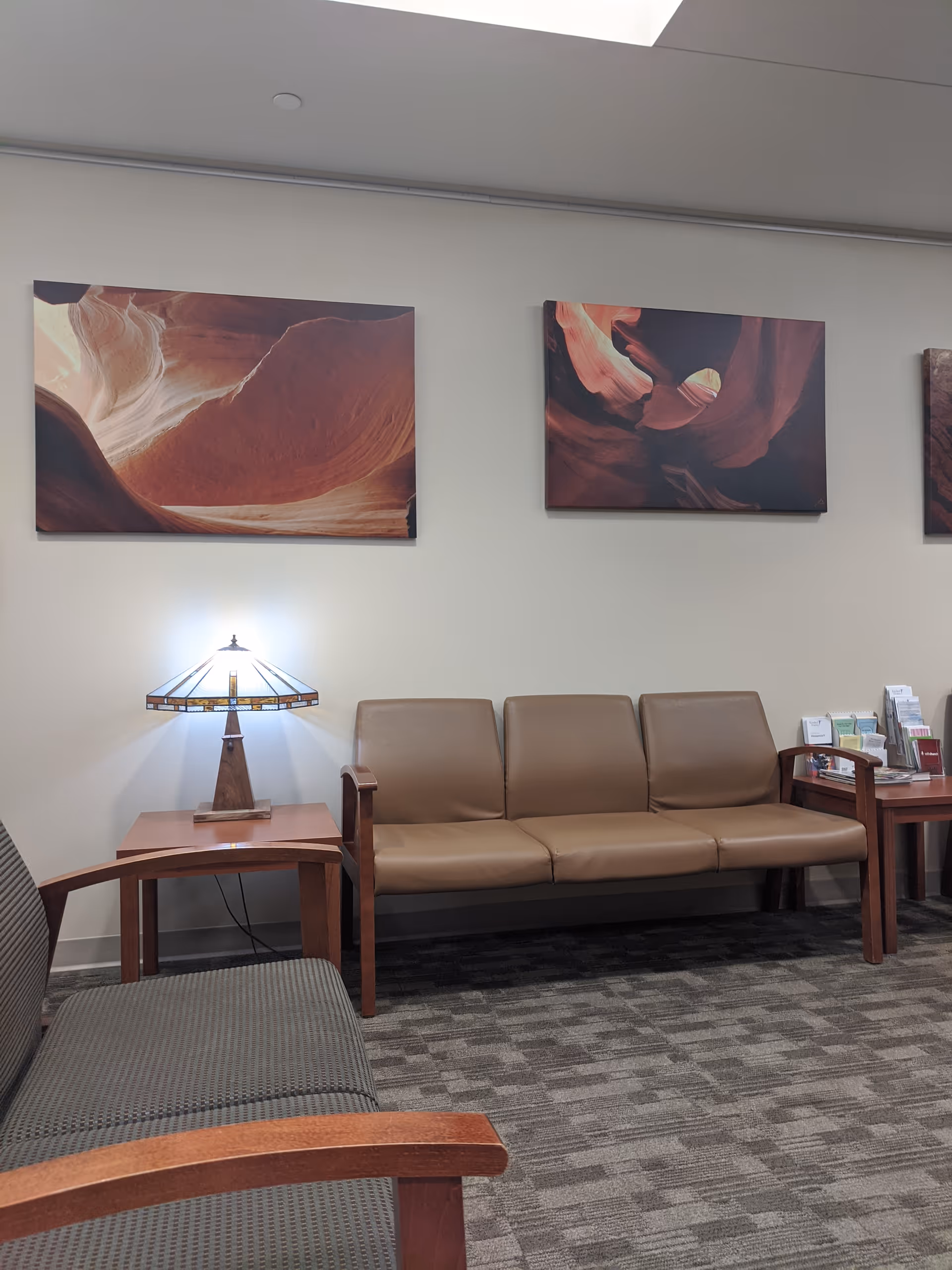 Lounge-style waiting area with tan leather sofas, wooden tables, a lamp, and wall art.
