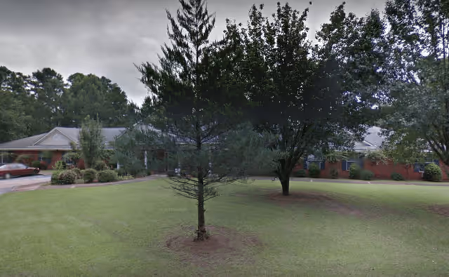 A single-story brick building with a gray roof surrounded by a well-maintained lawn and several trees, including a small pine tree in the foreground. There is a driveway with a parked car on the left side of the building.