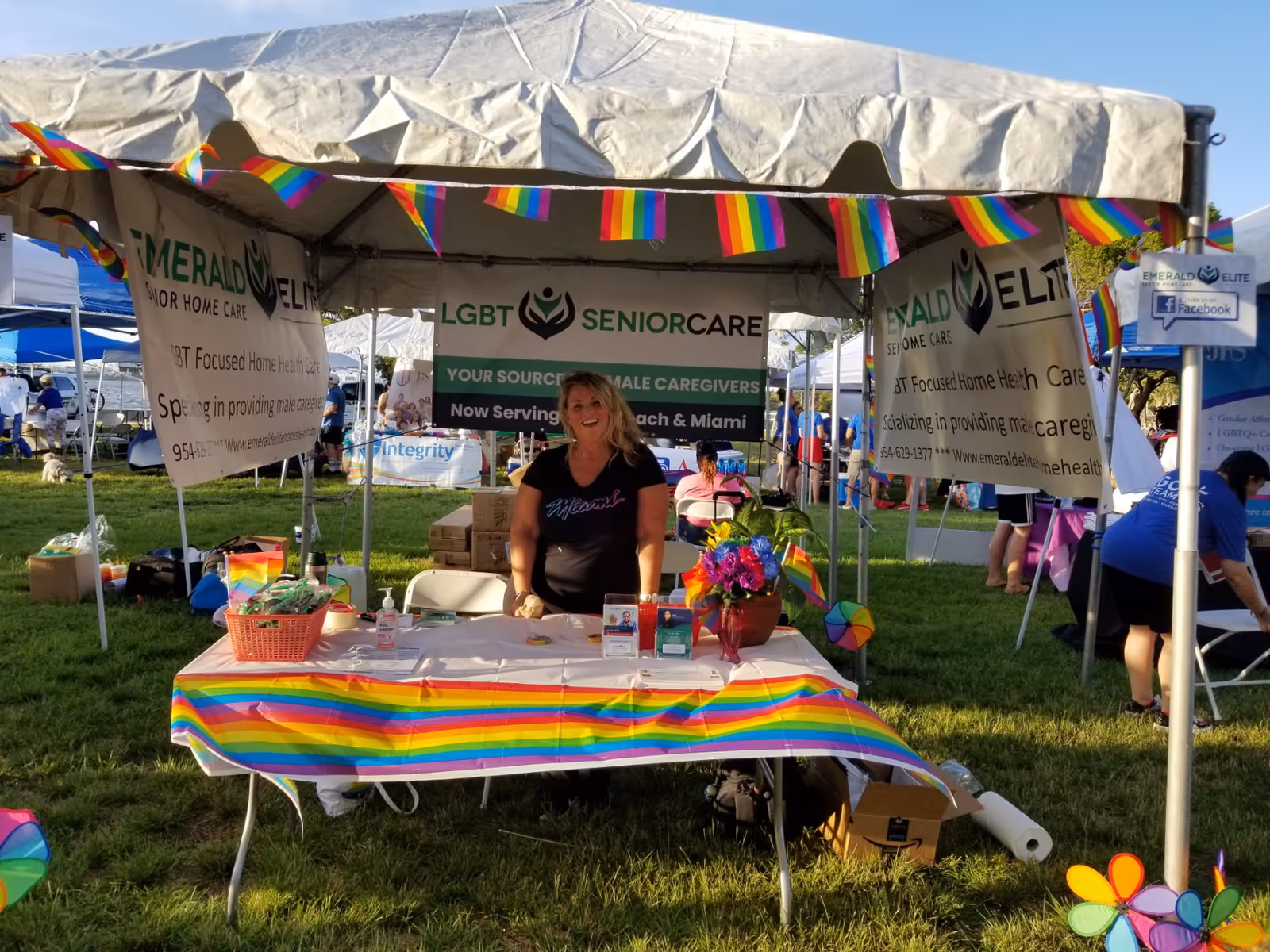 Woman at a rainbow-themed Emerald Senior Care booth outdoors at a community fair.