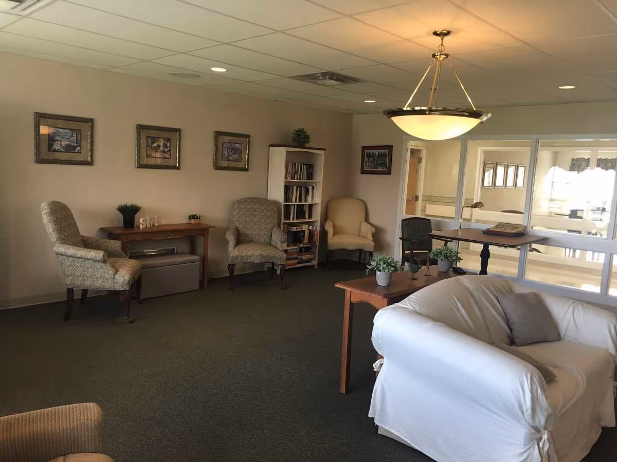 A communal sitting room with upholstered chairs, a white slipcovered sofa, tables, a bookshelf and framed pictures under a hanging ceiling light.