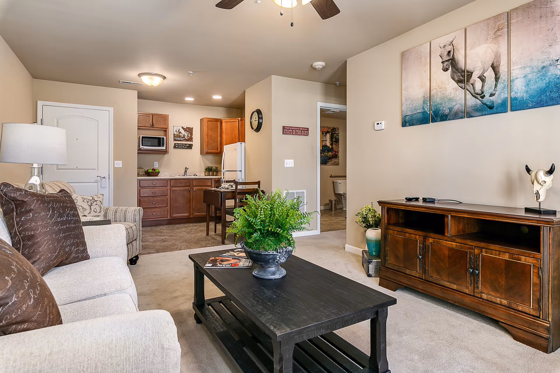 Open living room with a beige sofa and coffee table facing a wooden TV cabinet, with a kitchenette visible in the background.