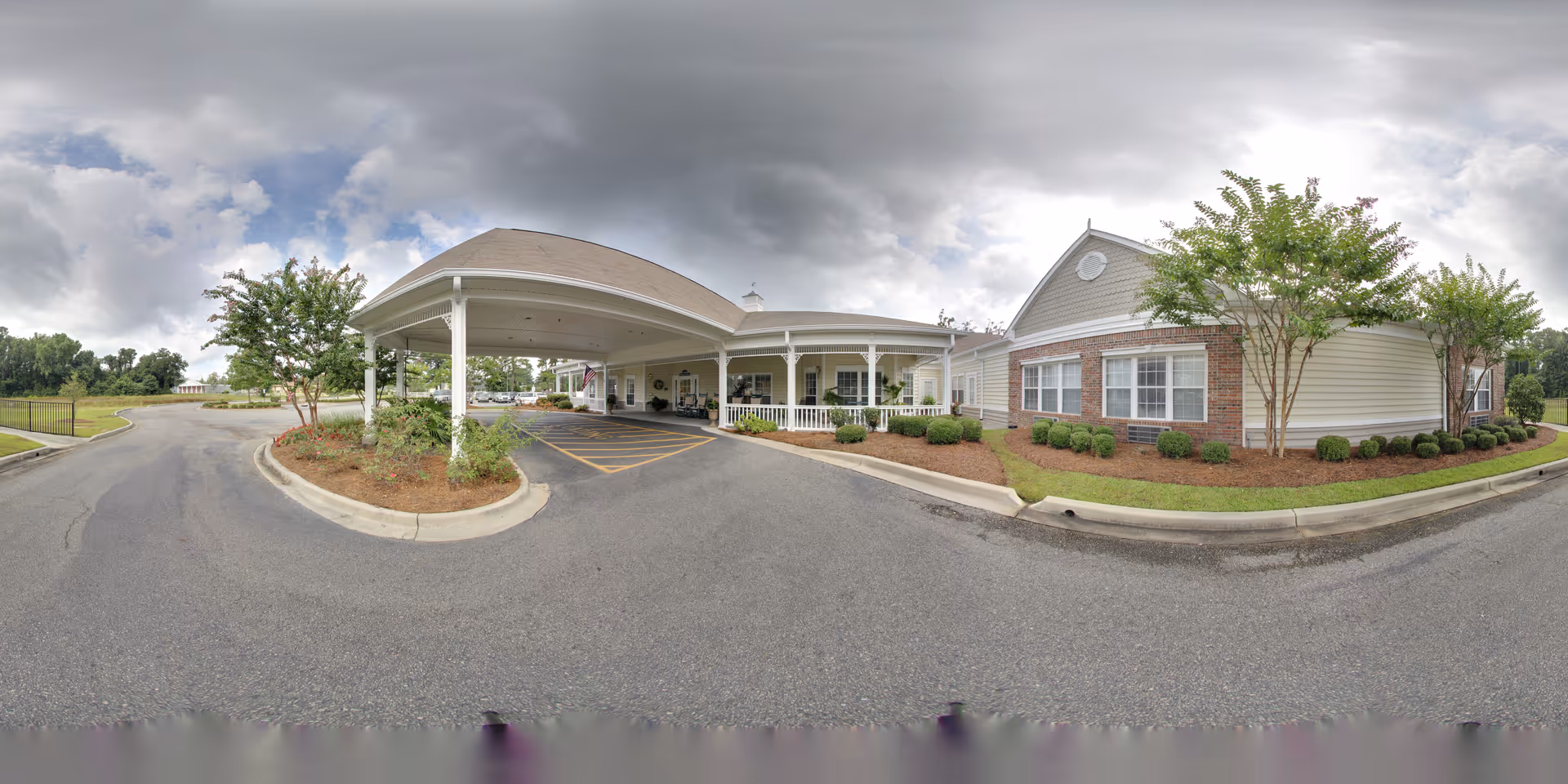 Exterior view of The Crossings of Georgetown senior living facility showing the main entrance with a covered drop-off area, landscaped greenery, and a paved driveway under a cloudy sky.