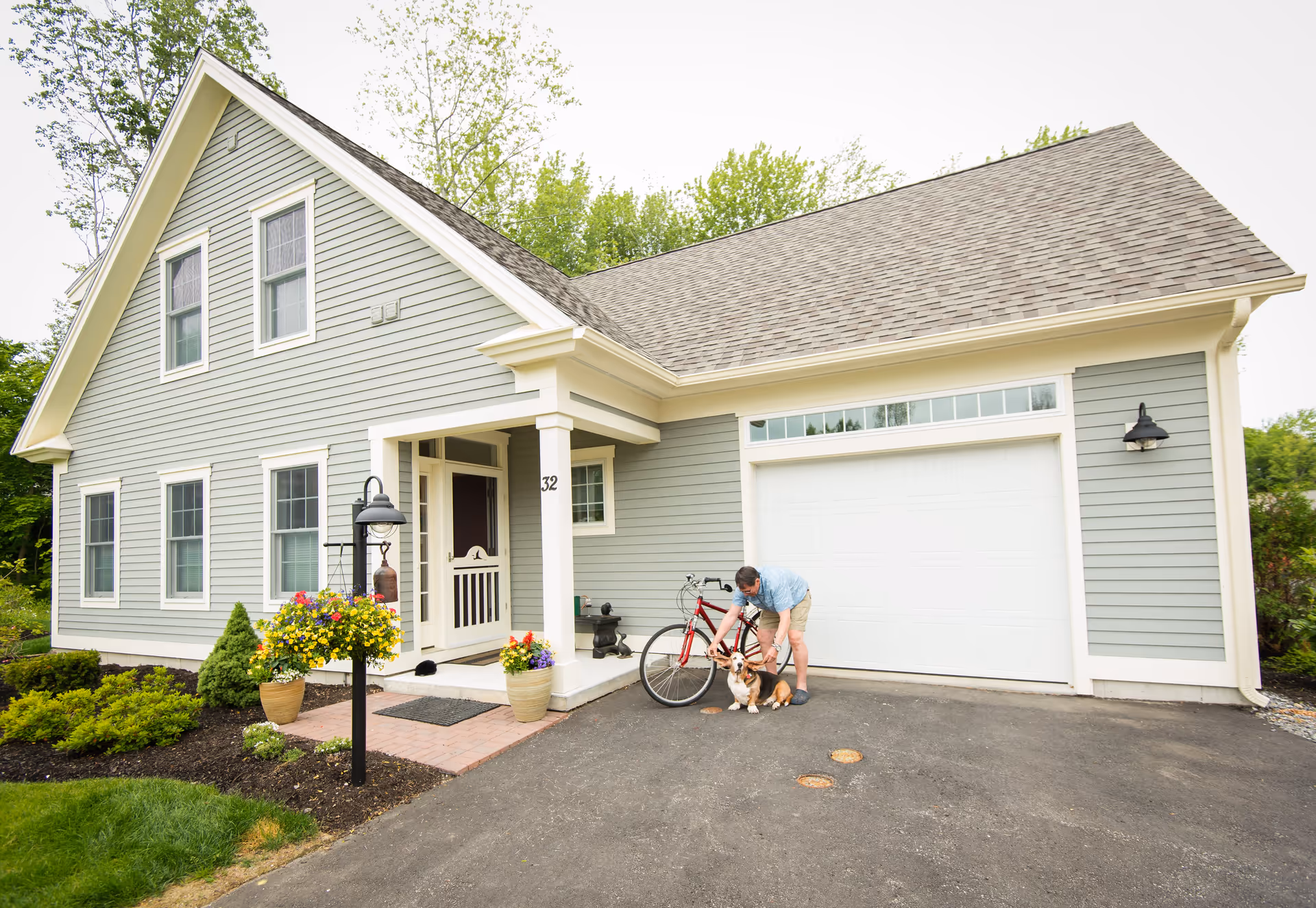 A light gray house with white trim and a large garage door. A person is bending down to pet a dog near a red bicycle parked in front of the garage. There are flower pots with colorful flowers near the entrance and a black lamp post with a hanging flower basket in the front yard.