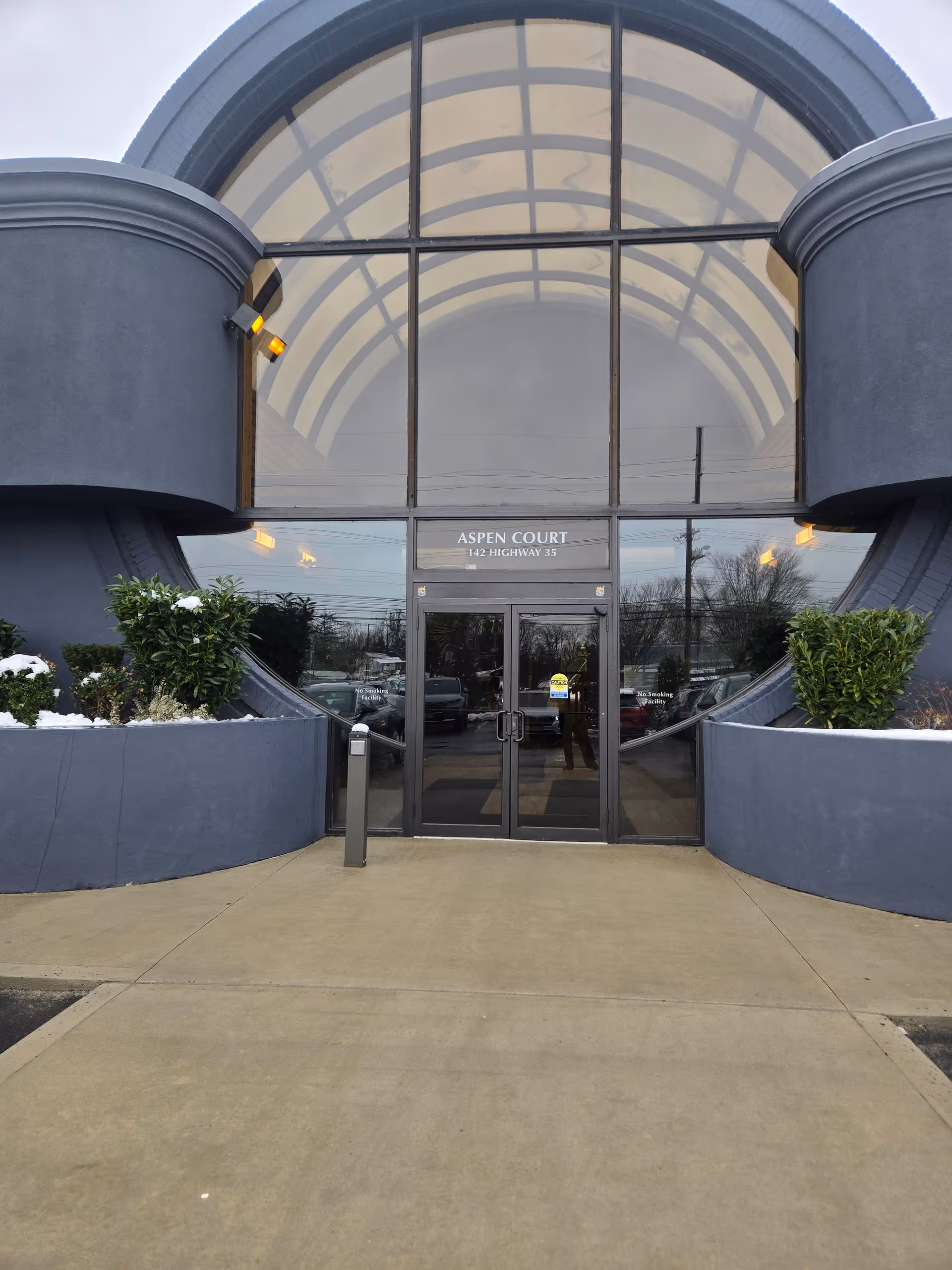 Modern building entrance with a large curved glass canopy and double doors, flanked by planters.