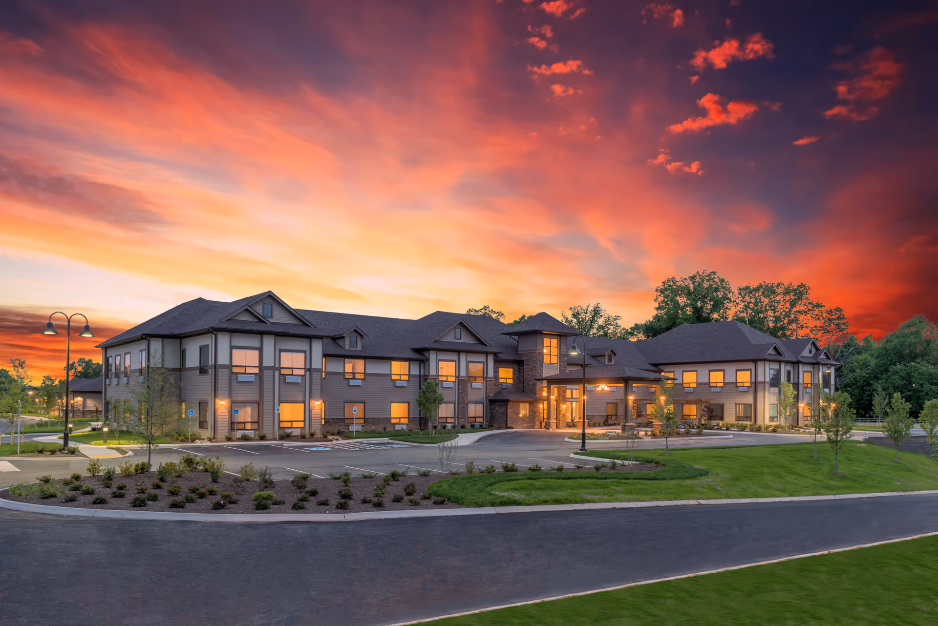 Exterior view of The Pointe at Lifespring Senior Living building at sunset with a vibrant orange and purple sky, well-lit windows, landscaped greenery, and a paved driveway in front.