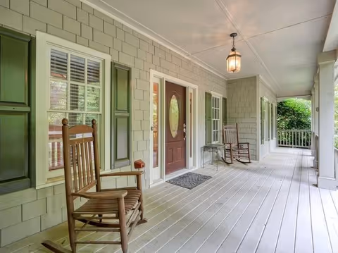 Covered front porch with rocking chairs, a red door, and green shutters along the house.