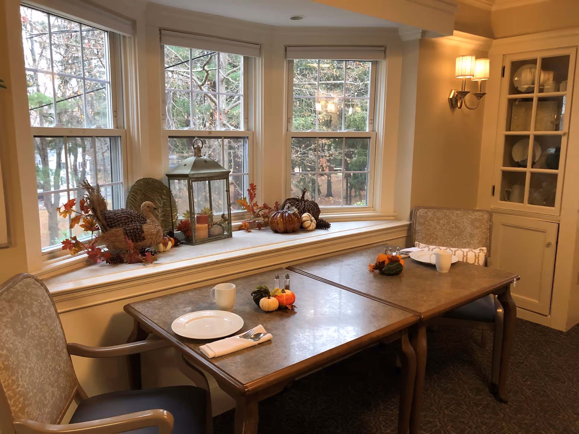 A cozy dining area with a table set for two, featuring plates, cups, and napkins. The table is decorated with small pumpkins and gourds. Behind the table is a large bay window with autumn-themed decorations including a turkey figurine, a lantern, and various fall leaves and pumpkins. The room has warm lighting with a wall sconce and a built-in cabinet displaying dishes.