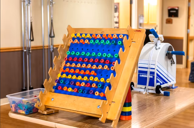 A wooden pegboard game with colorful pegs arranged in rows on a table, next to a white and blue exercise machine in a room with wooden floors and beige walls.