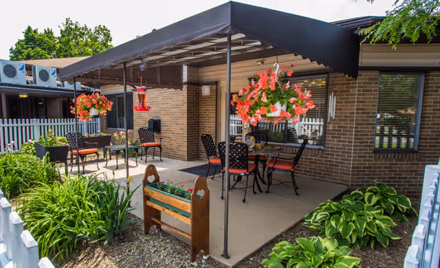 Outdoor patio area at Grove Manor with a black canopy, several tables and chairs with red cushions, hanging flower baskets with pink flowers, green plants and shrubs surrounding the patio, and a white picket fence in the background.