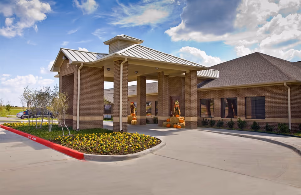 Exterior view of a brick building with a covered entrance supported by brick columns. The driveway curves around a landscaped area with flowers and small trees. There are autumn decorations with pumpkins and hay bales near the entrance. The sky is partly cloudy.