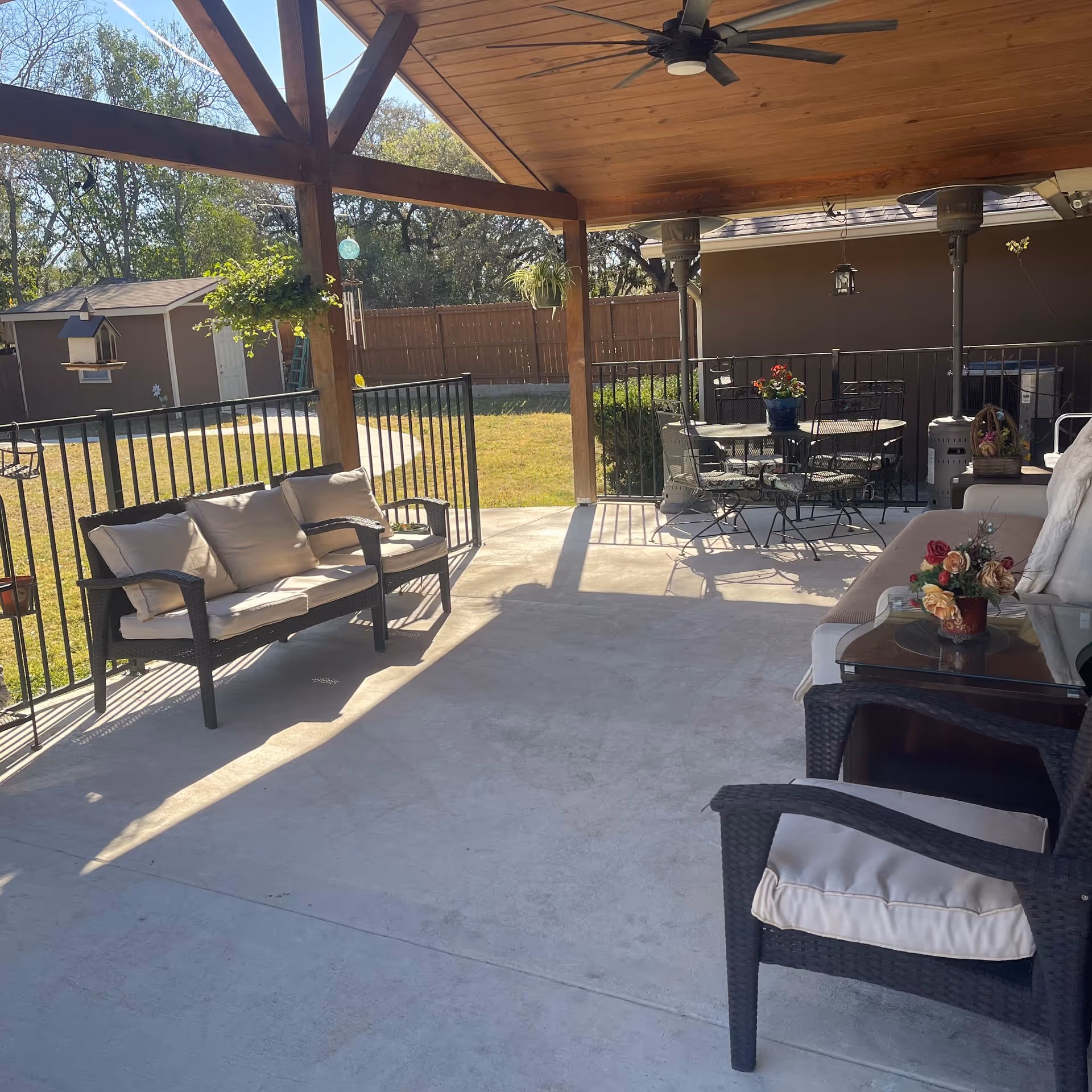 Covered outdoor patio area with cushioned wicker seating including a loveseat and chairs, a glass-top table with a flower arrangement, and a round metal dining table with chairs. The patio has a wooden ceiling with a ceiling fan and is surrounded by a black metal fence. In the background, there is a grassy yard, a wooden fence, and a small shed.