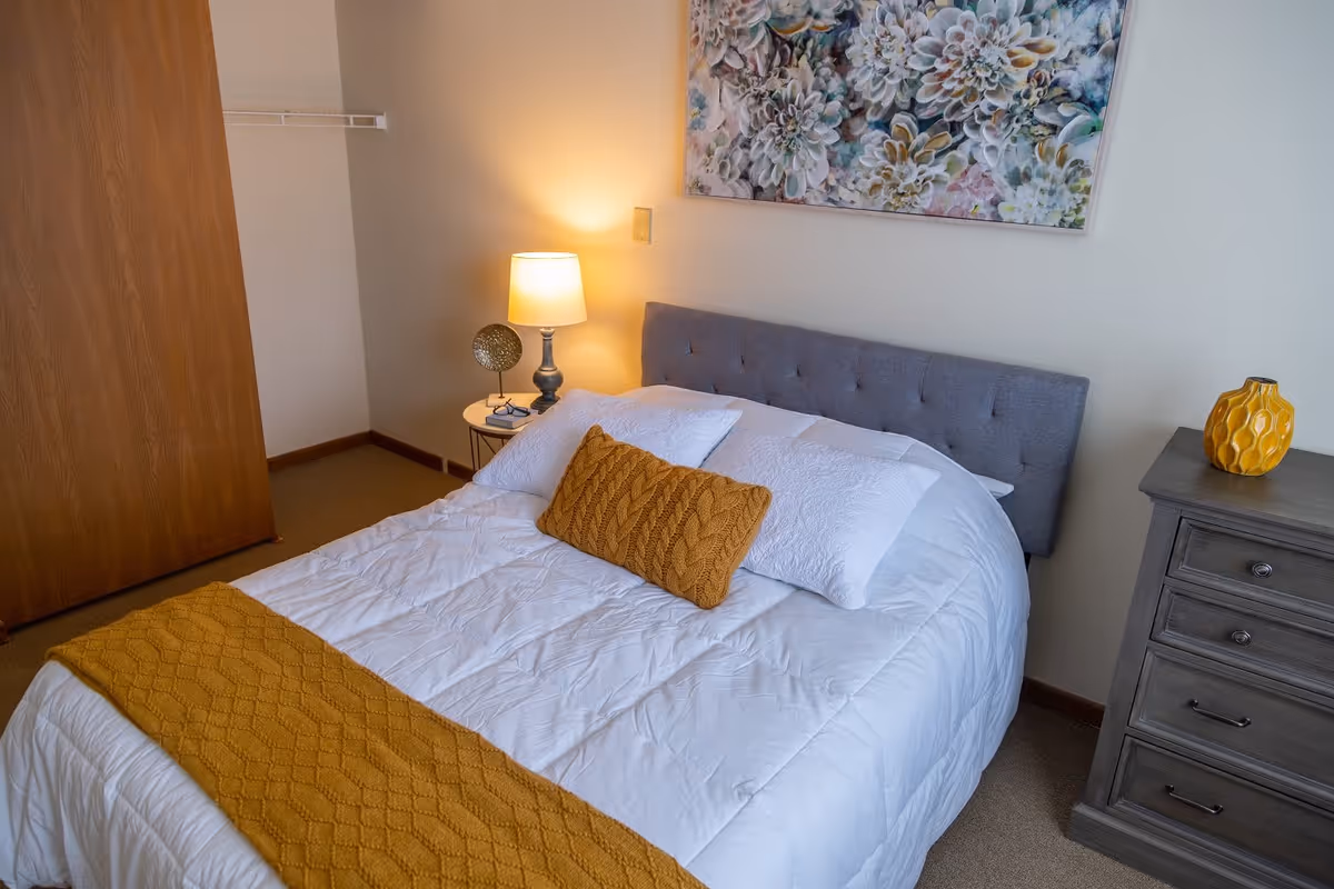 A neatly made bed with white bedding and mustard yellow knitted throw blanket and pillow in a bedroom. There is a gray upholstered headboard, a wooden dresser with a yellow vase, a small round bedside table with a lamp and decorative item, and a floral painting on the wall above the bed. A wooden closet door is visible on the left side.