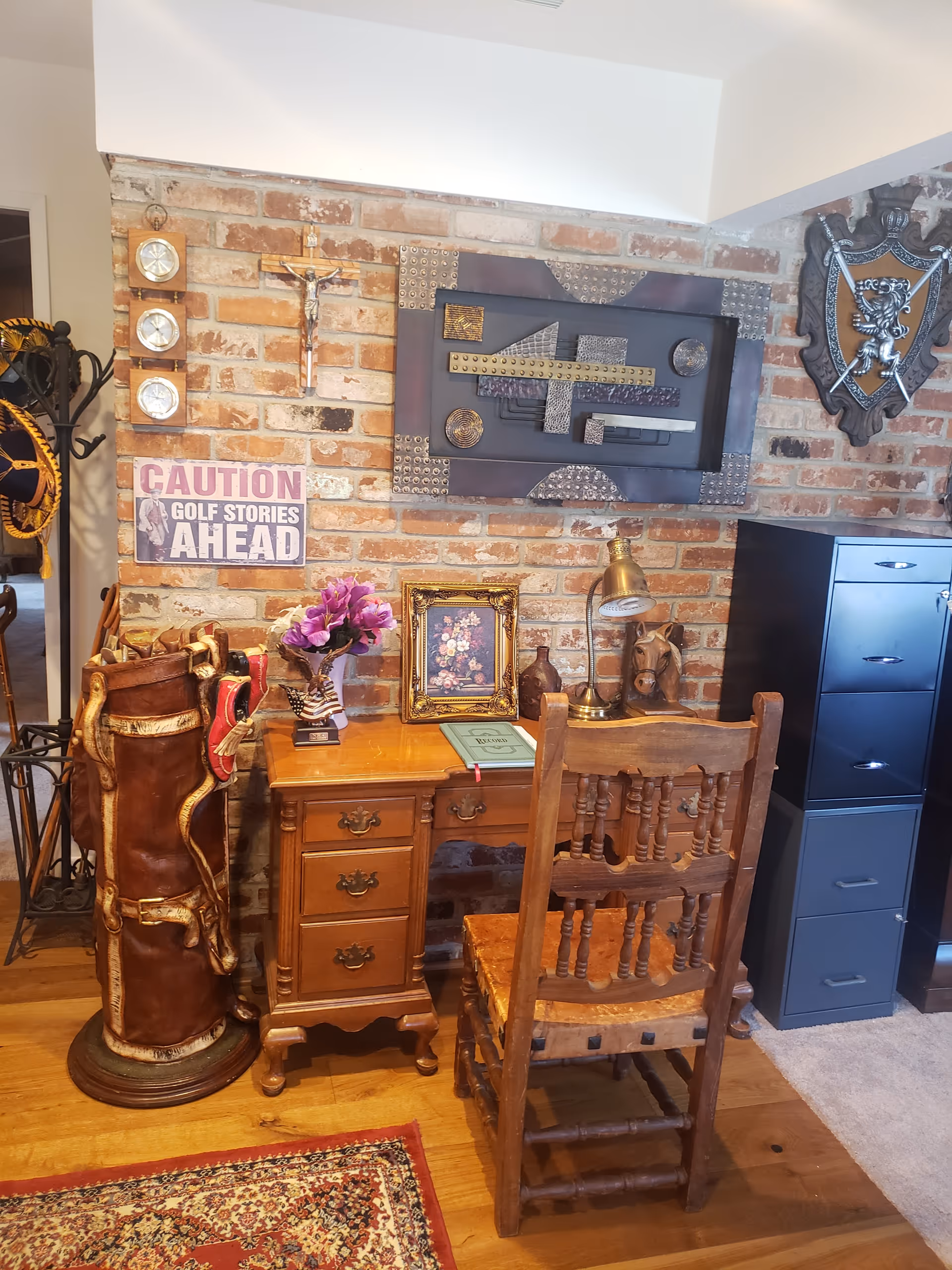 A cozy interior corner featuring a wooden desk with a matching wooden chair. On the desk are a framed floral picture, a vase with purple flowers, a desk lamp, and a book. Behind the desk is a brick wall adorned with various decorations including a crucifix, a sign that reads 'CAUTION GOLF STORIES AHEAD', a decorative shield, and an abstract metal wall art piece. To the left of the desk is a vintage golf bag with clubs, and to the right is a black filing cabinet. The floor is wooden with a patterned rug partially visible.