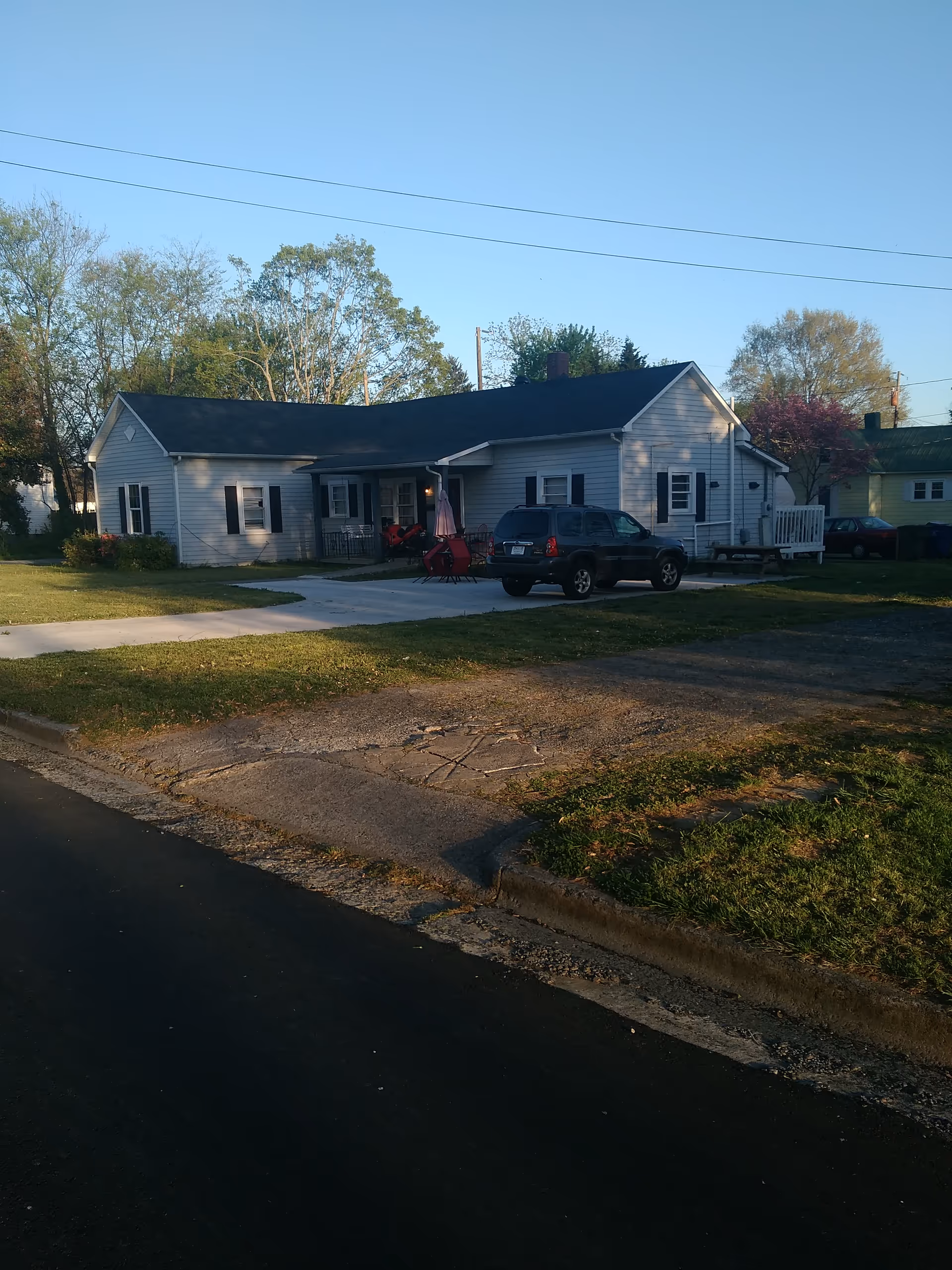 Single-story residential facility exterior with a driveway, parked SUV, and a small front porch seating area.