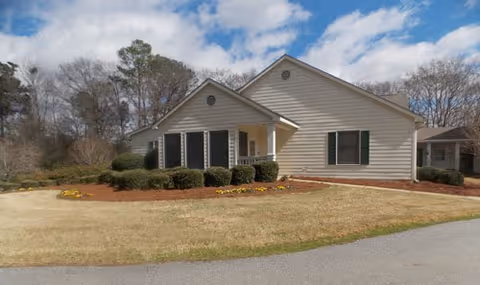 Single-story assisted living facility with beige siding, trimmed shrubs, and a paved front driveway.