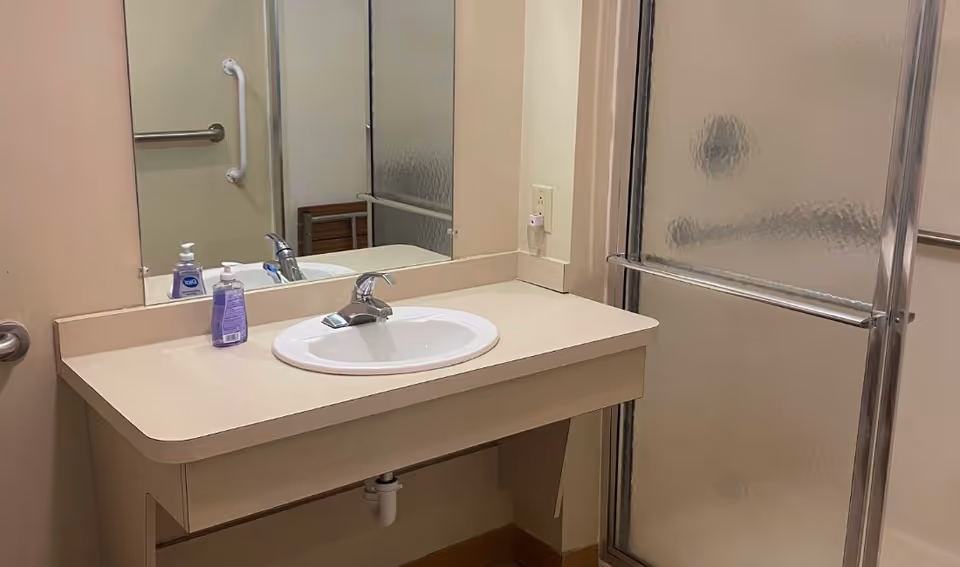 Bathroom with sink, mirror, soap dispensers, and frosted-glass shower in a facility.