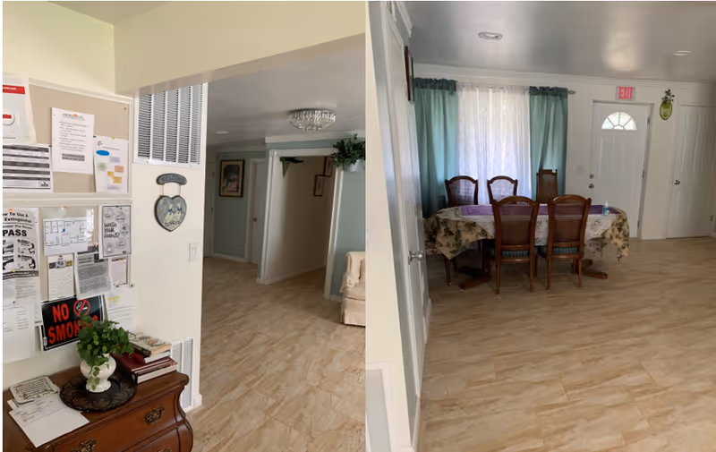Interior view of a senior living facility showing a hallway with a bulletin board filled with notices and a small table with a plant and books on the left. The hallway leads to a dining area with a table covered with a floral tablecloth and six wooden chairs. There is a window with sheer curtains and green drapes, and a white door with an exit sign above it.