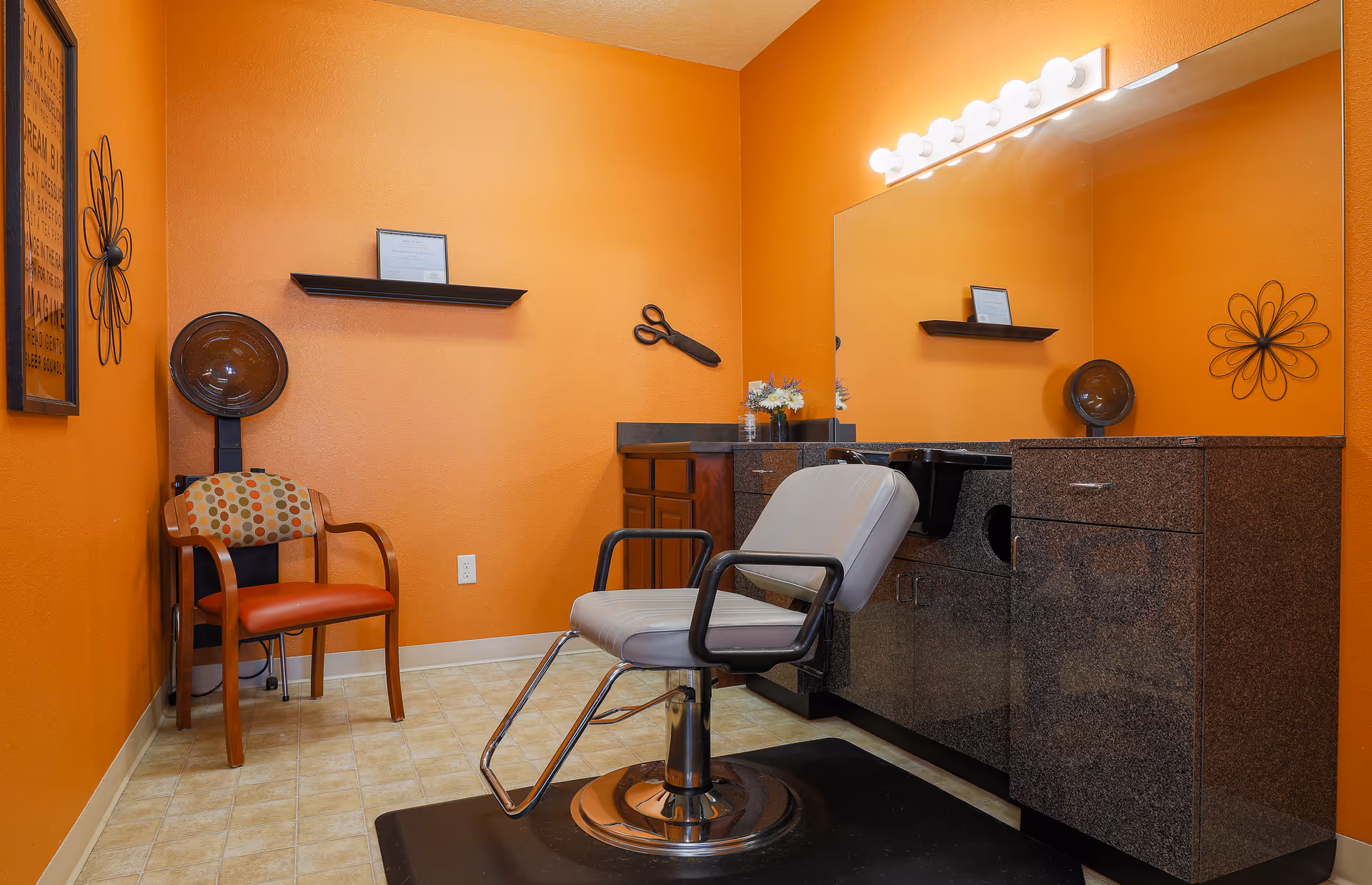 Interior of a hair salon room with orange walls, a salon chair in front of a large mirror, a hair dryer hood next to a wooden chair with a patterned cushion, and a countertop with cabinets and a sink.