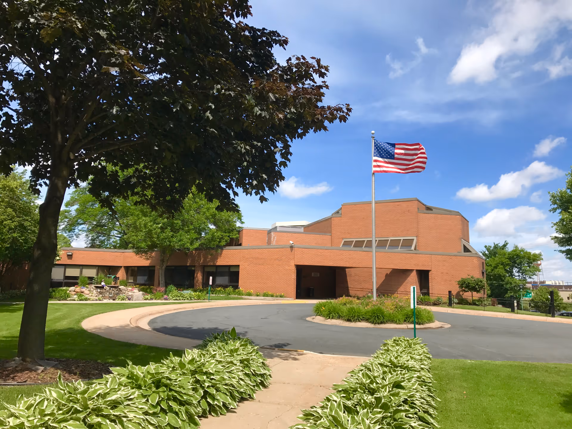 Brick hospice building with circular driveway, flagpole, and manicured landscaping under a blue sky.