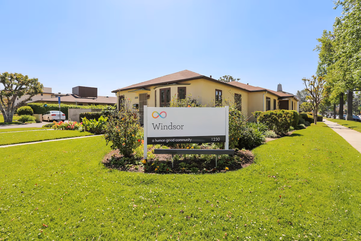 Exterior view of a single-story building with a brown roof and beige walls, surrounded by green grass, bushes, and trees under a clear blue sky. In front of the building is a white sign with the text 'Windsor a human good community 1230'. A sidewalk runs along the right side of the image.