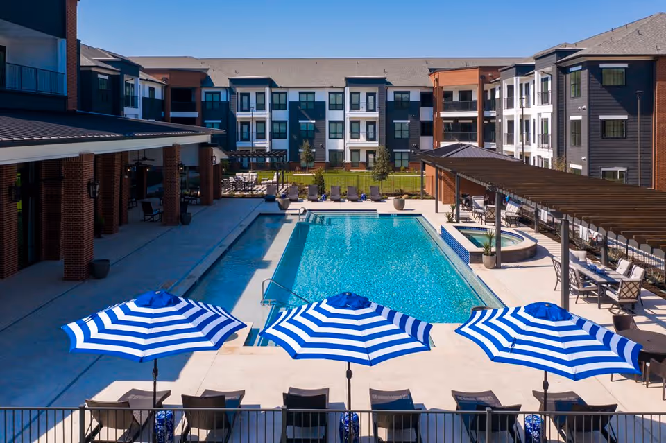 Outdoor swimming pool area at Northstar Active Adult 55 Plus facility featuring a rectangular pool, lounge chairs, three blue and white striped umbrellas, a hot tub, and a covered seating area with tables and chairs. The pool is surrounded by a multi-story residential building under a clear blue sky.