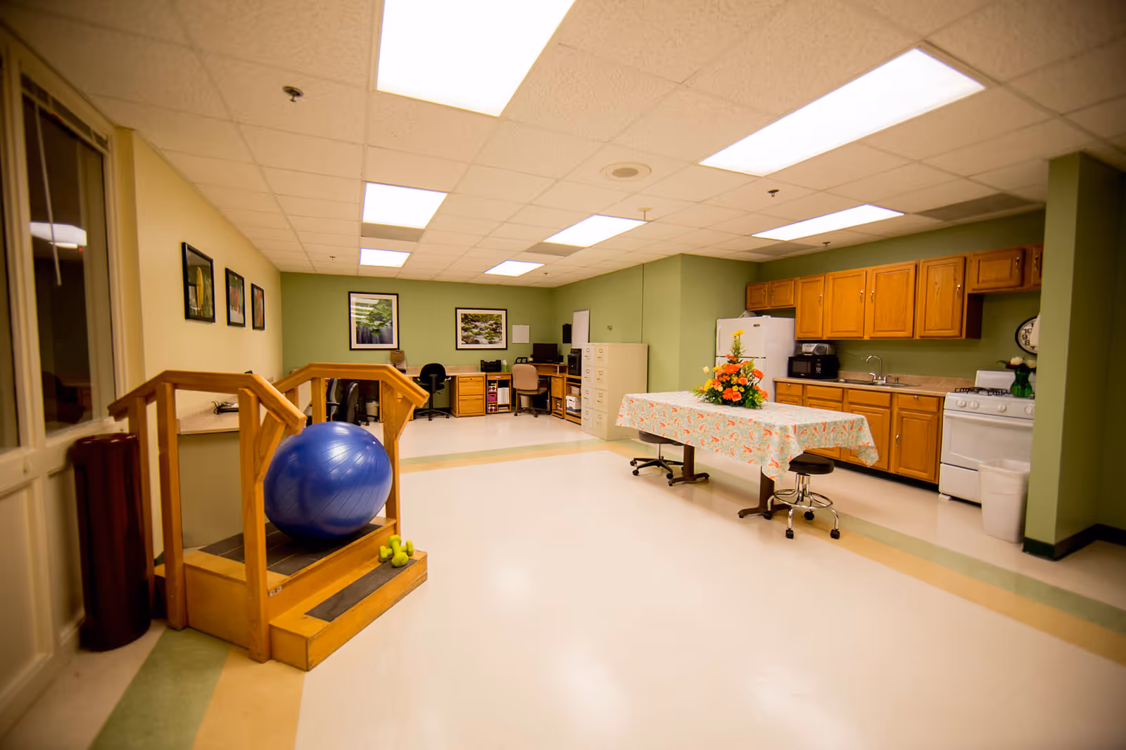 Interior common area of a senior living facility featuring a kitchen, table, exercise ball, and computers.