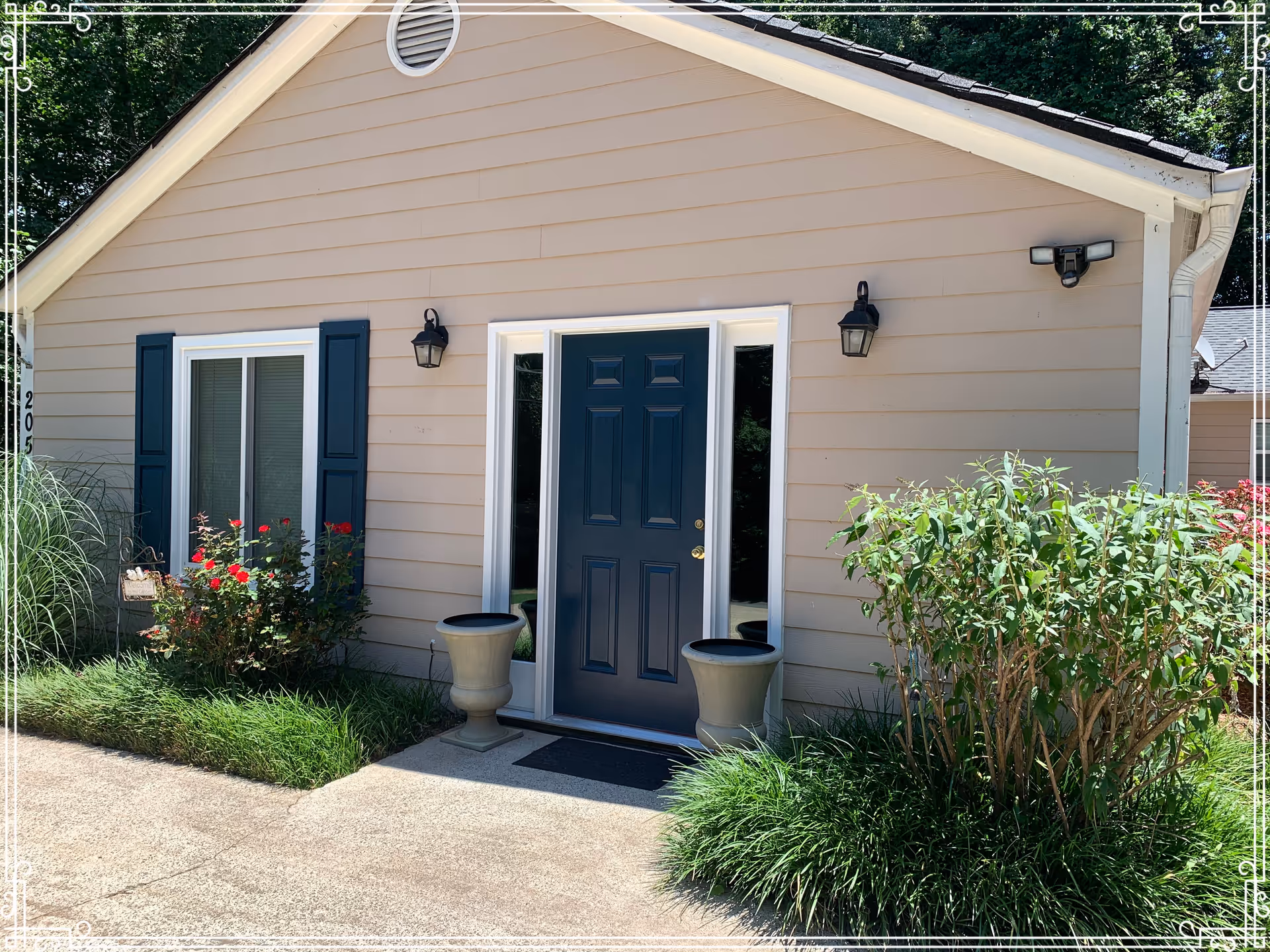 Exterior view of a single-story building with beige siding and a dark blue front door flanked by two narrow windows. There are two black wall-mounted lantern lights on either side of the door. The building has a small garden area with green shrubs and red flowers near the entrance, and a concrete pathway leading to the door.