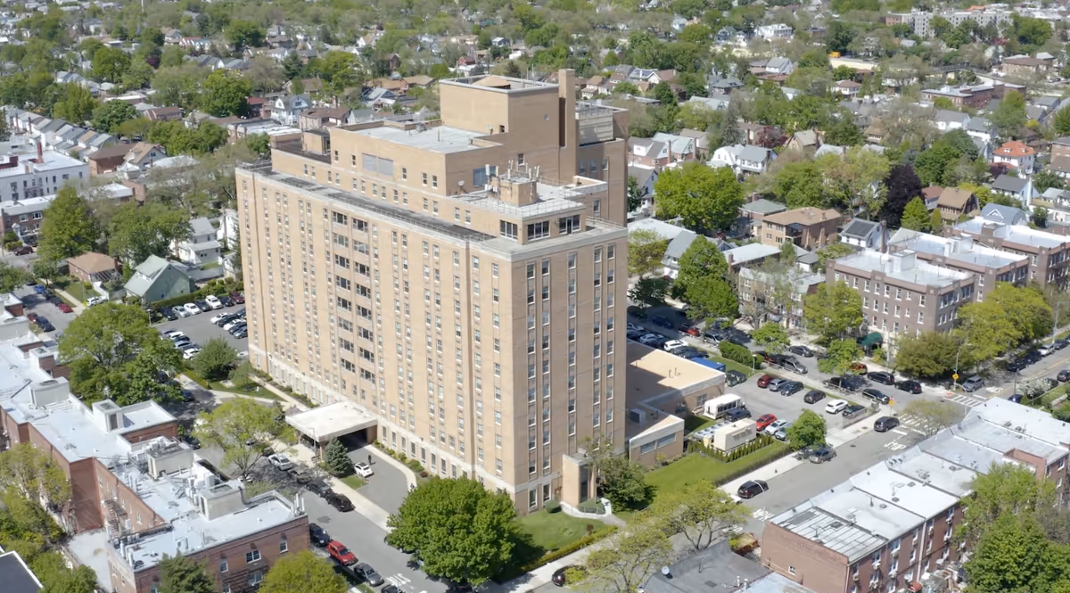 Aerial view of a large multi-story brick building surrounded by trees and residential houses, identified as Ozanam Hall of Queens Nursing Home.